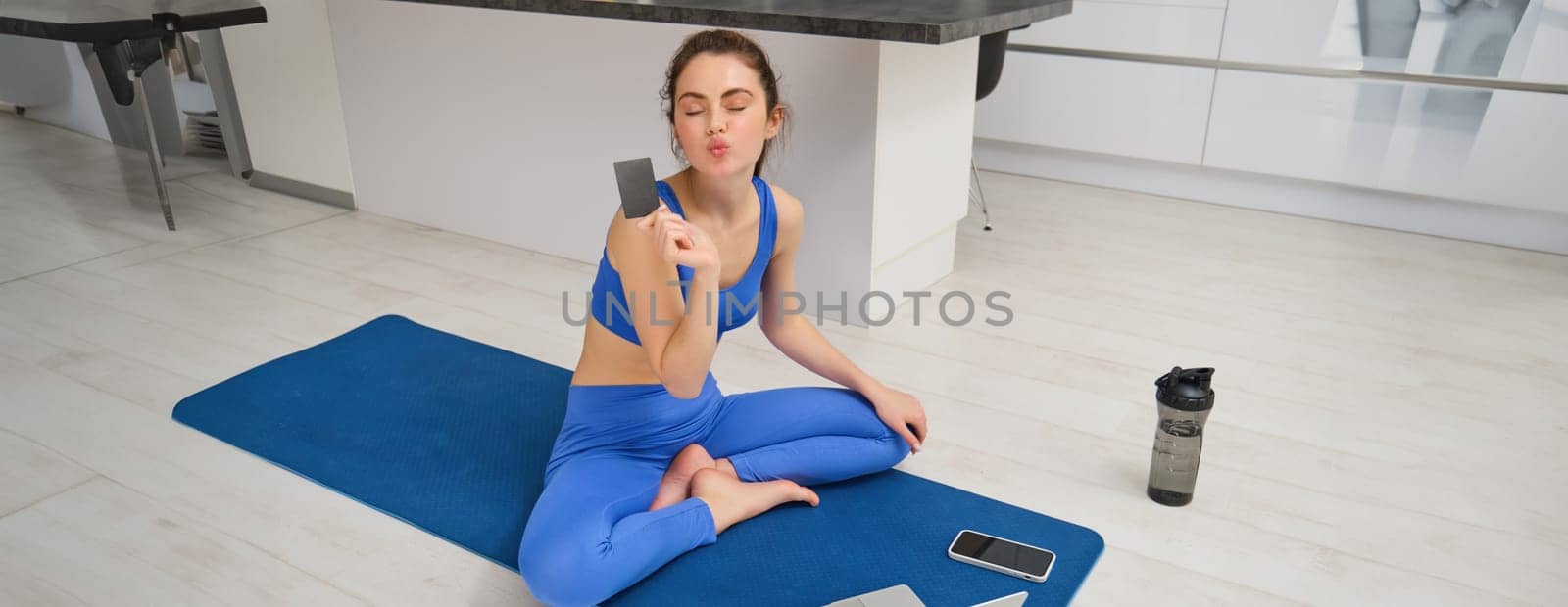 Image of smiling young woman, holding credit card, sitting on fitness mat with laptop, buying sport app subscription, workout video tutorials, doing exercises at home.