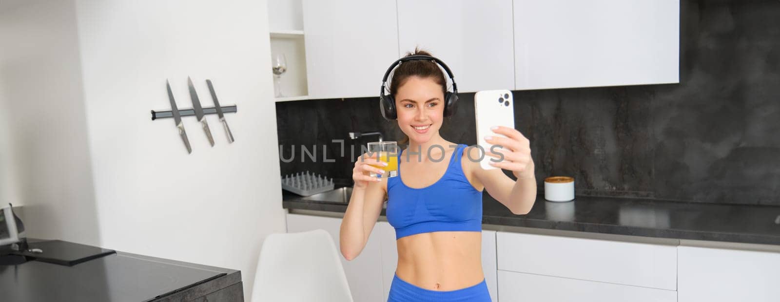 Portrait of young lifestyle, fitness blogger, woman with orange juice, takes selfie on smartphone camera, posing in activewear and wireless headphones.