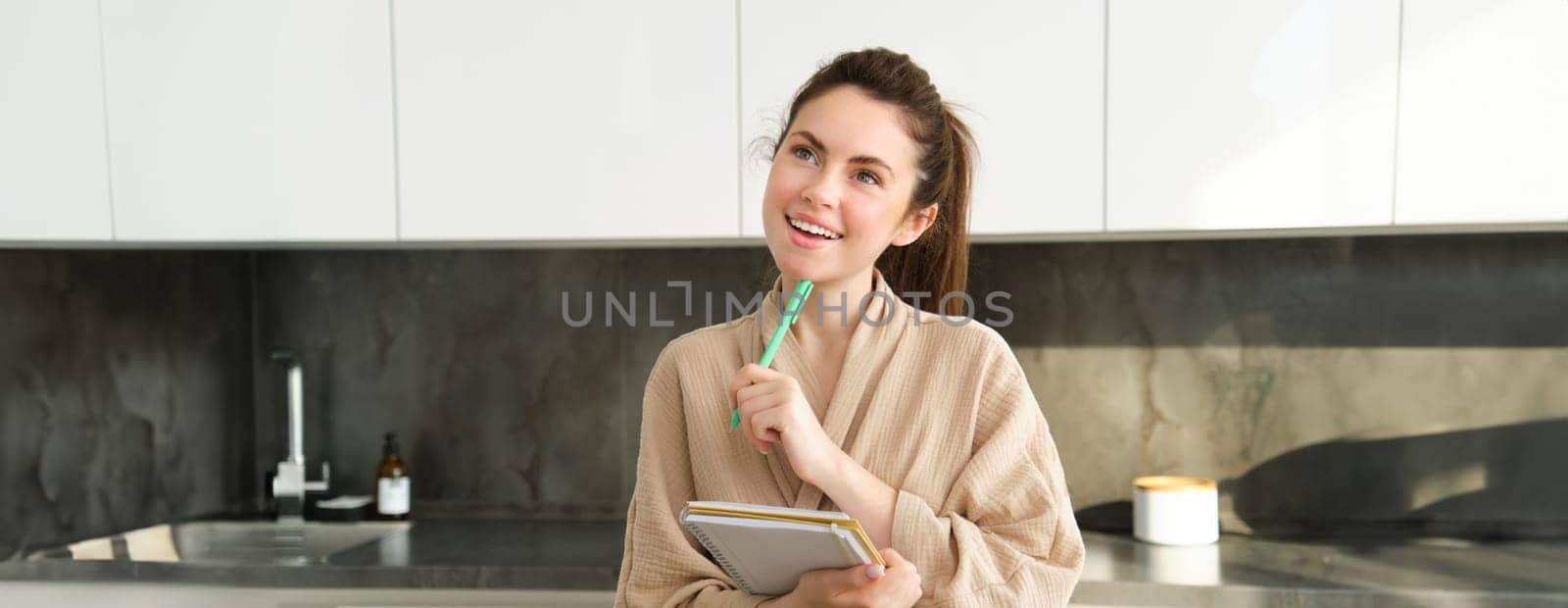 Portrait of woman preparing grocery list, writing down food ideas, creating new recipe in the kitchen, thinking of meal list for dinner, standing in bathrobe by Benzoix