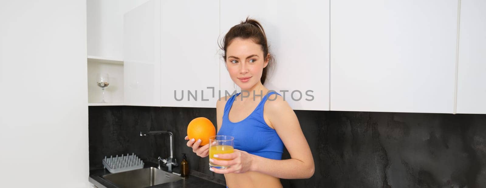 Indoor shot of young brunette woman in sportswear, drinking orange juice, holding fruit in hand, posing in kitchen.