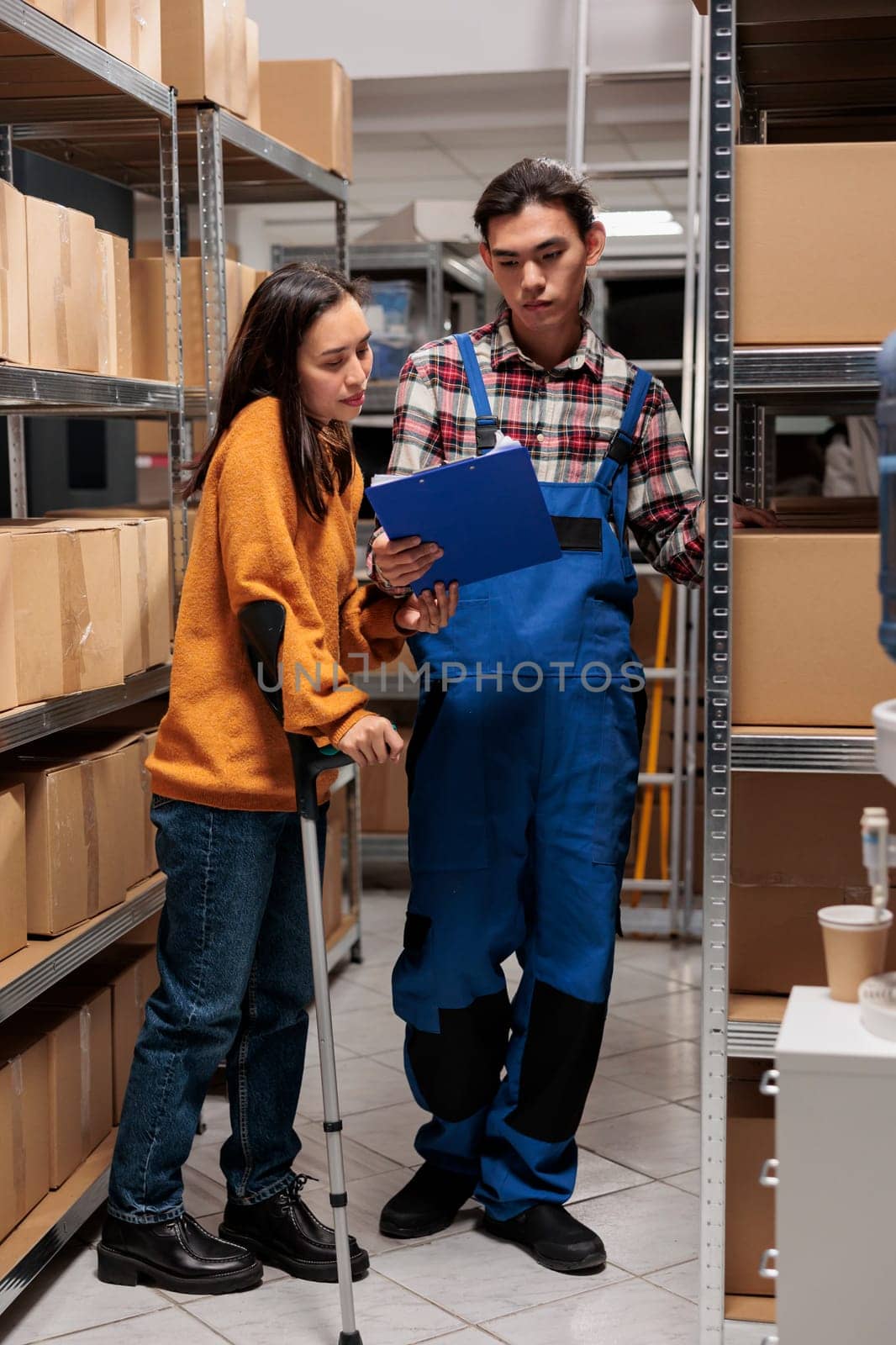 Storehouse asian employees managing inventory optimization in storage room. Warehouse logistics manager on crutches and young man pakcage handler analyzing distribution report on clipboard