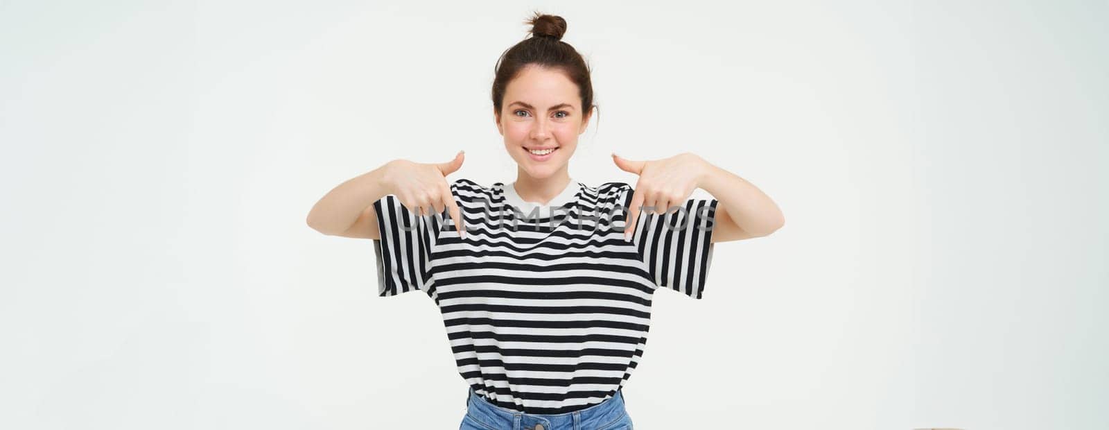 Portrait of beautiful, smiling young female model, showing advertisement, pointing fingers down, standing over white background.