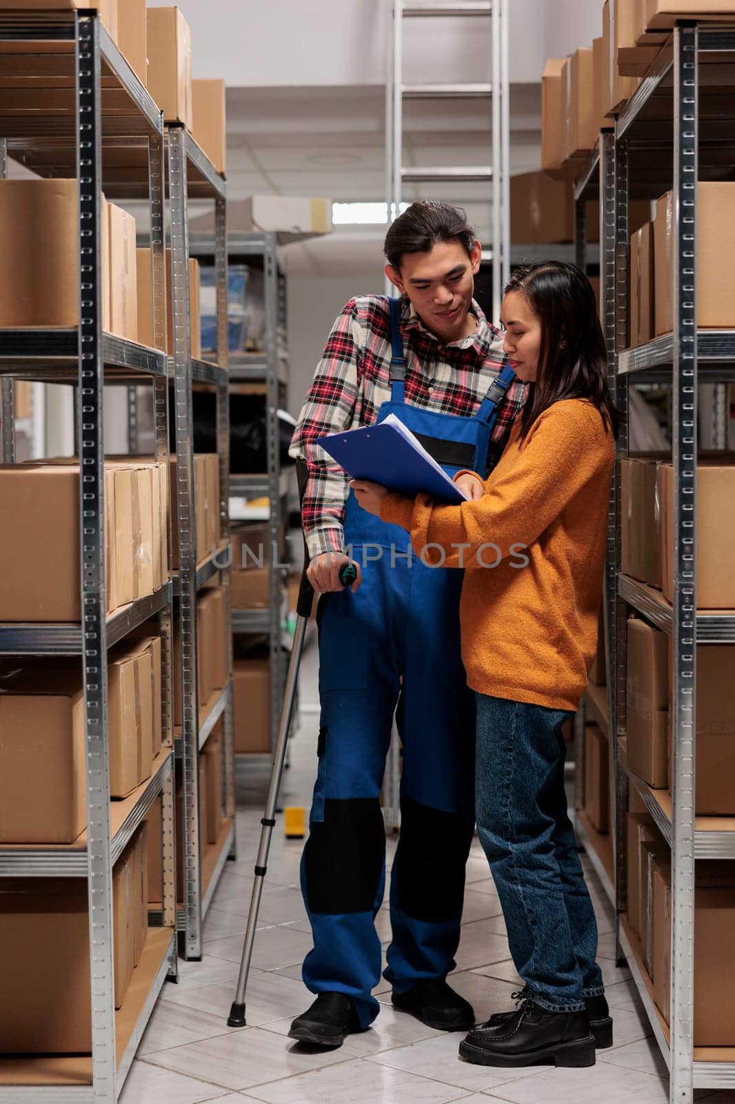 Warehouse manager explaining stock tracking system to package handler on crutches. Storehouse employees standing in storage room aisle and analyzing orders checklist on clipboard