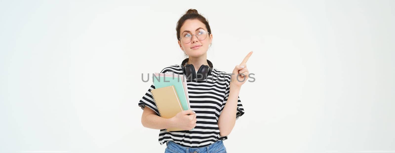 Image of smart young woman in glasses, student carries her notebooks and planners, points left with thinking face, making choice, deciding on smth, white background.