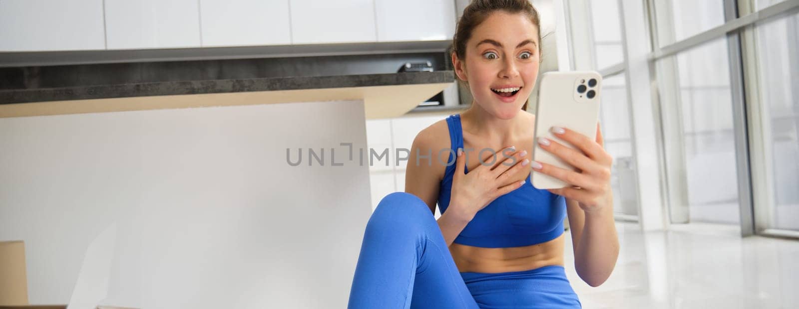 Close up shot of young happy woman, doing sports, workout at home, checking her phone, looking amazed at smartphone screen. Copy space