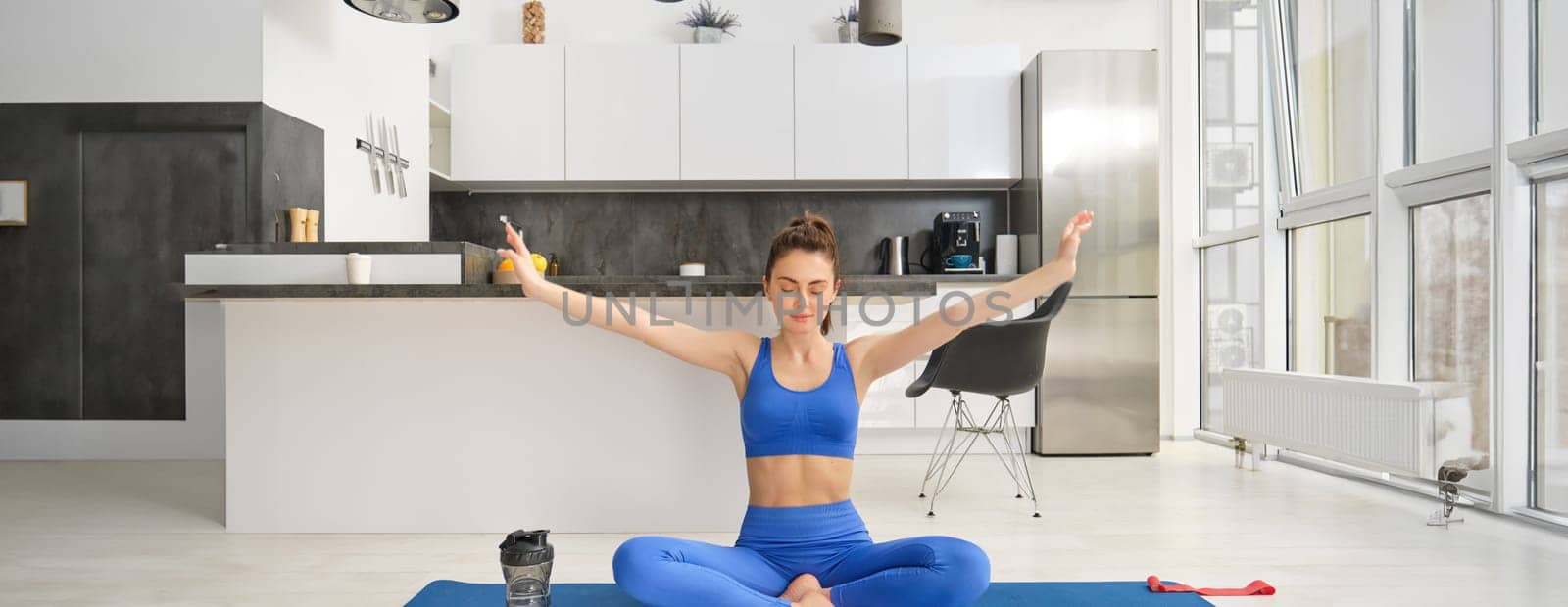 Young woman doing sports, meditating on yoga mat at home, holds hands sideways and breathing, relaxing mindful exercises.