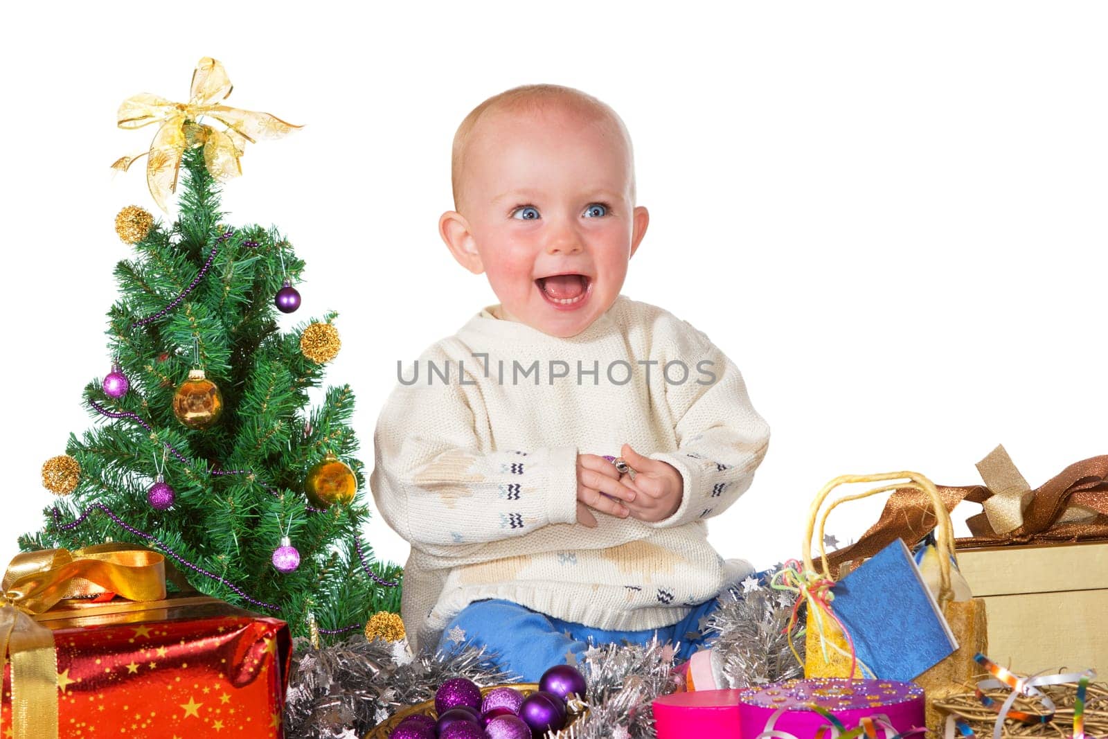 Adorable laughing baby sitting on the floor surrounded by a Christmas tree, gifts and decorations
