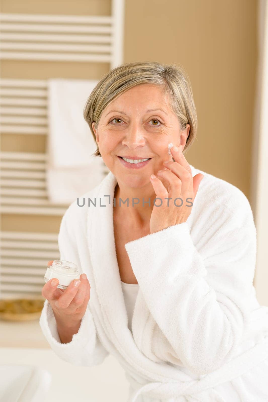 Senior woman hold anti-wrinkles cream in bathroom looking at camera