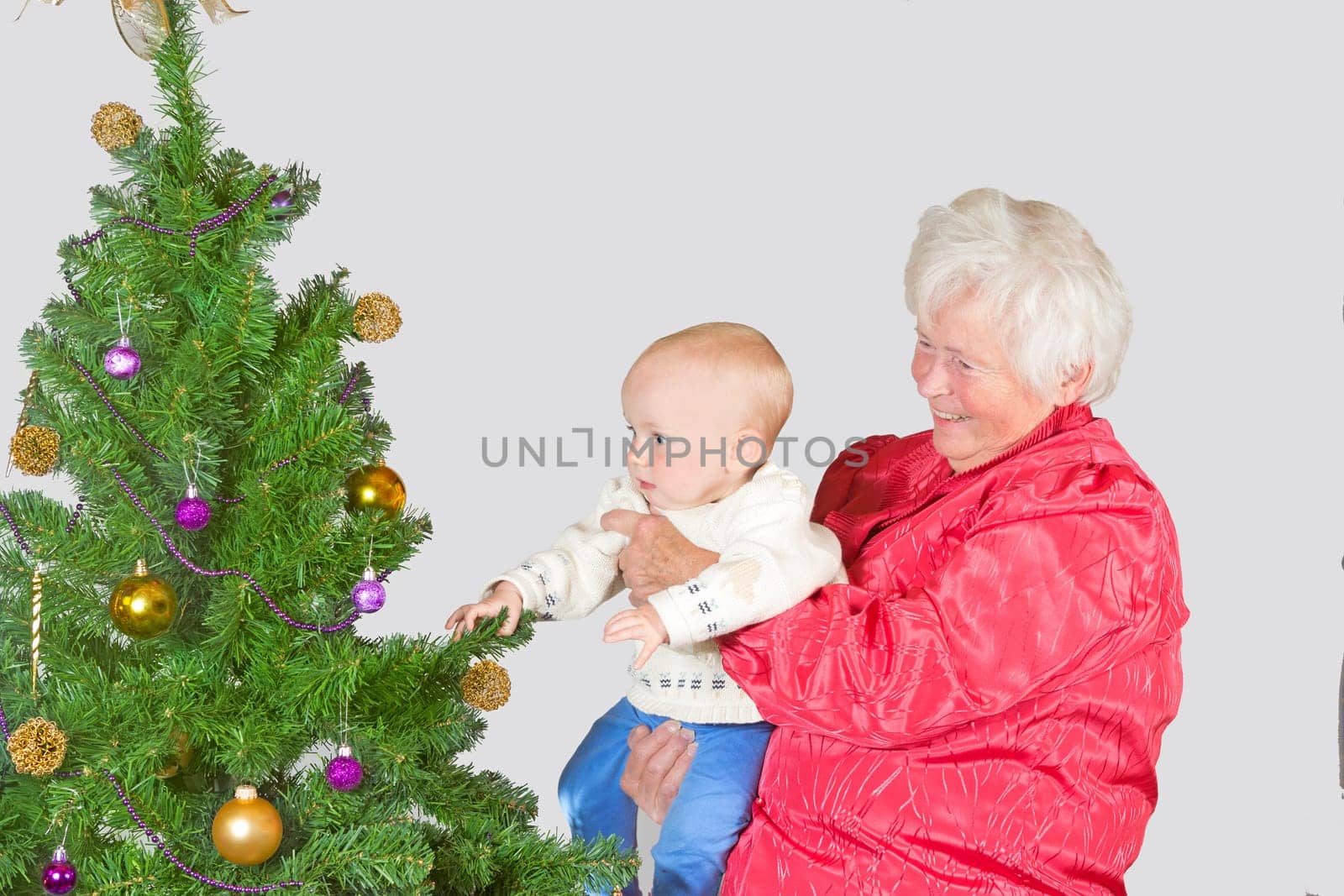 A happy smiling grandmother holds a cute young baby in her arms as they inspect a decorated Christmas tree