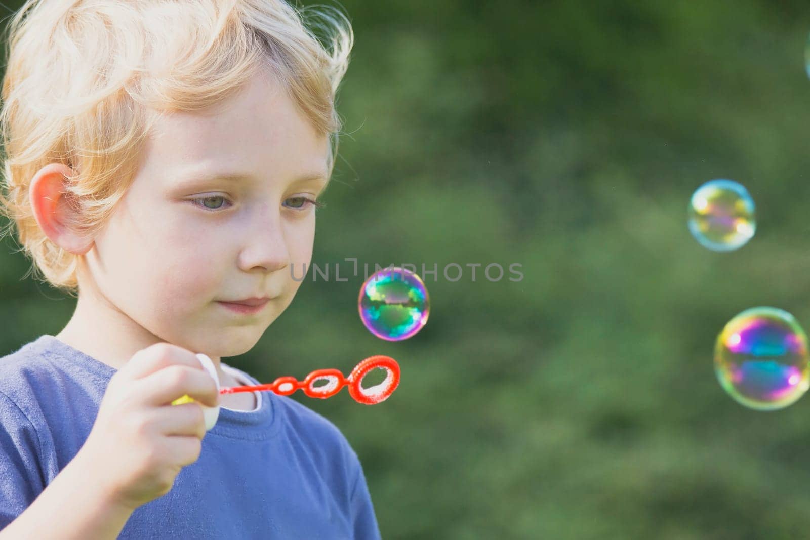 Blond boy in blue shirt blows  bubbles in the garden.