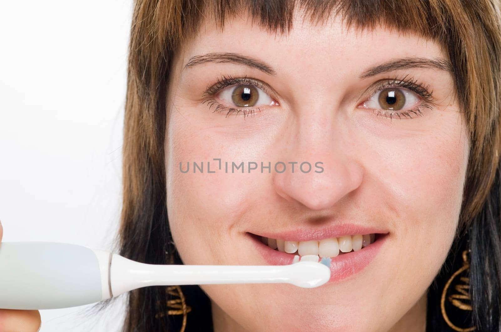Frontal head shot of young brunette woman with electric toothbrush smiling looking at camera against white background