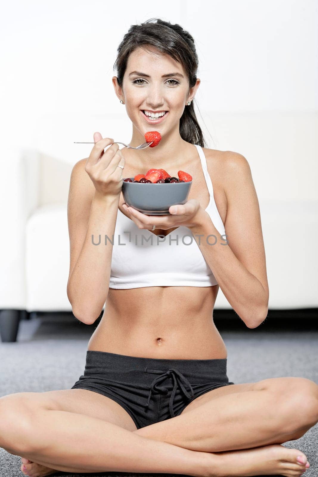 beautiful young woman in fitness clothes eating fresh fruit from a bowl at home