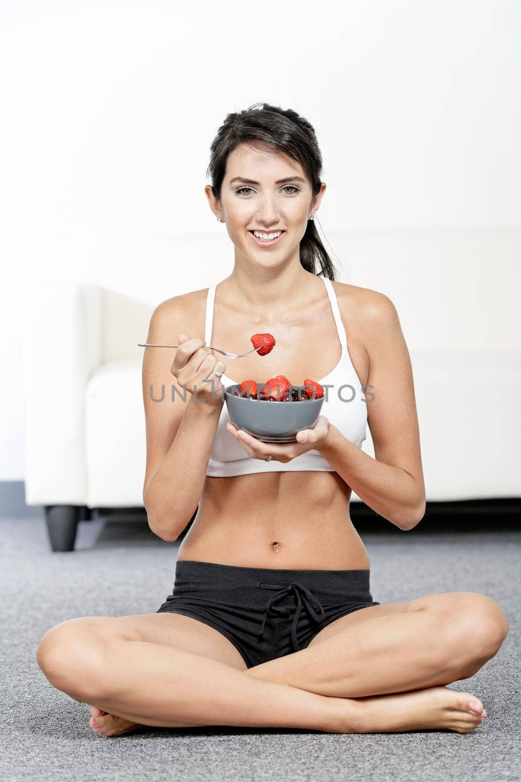 beautiful young woman in fitness clothes eating fresh fruit from a bowl at home