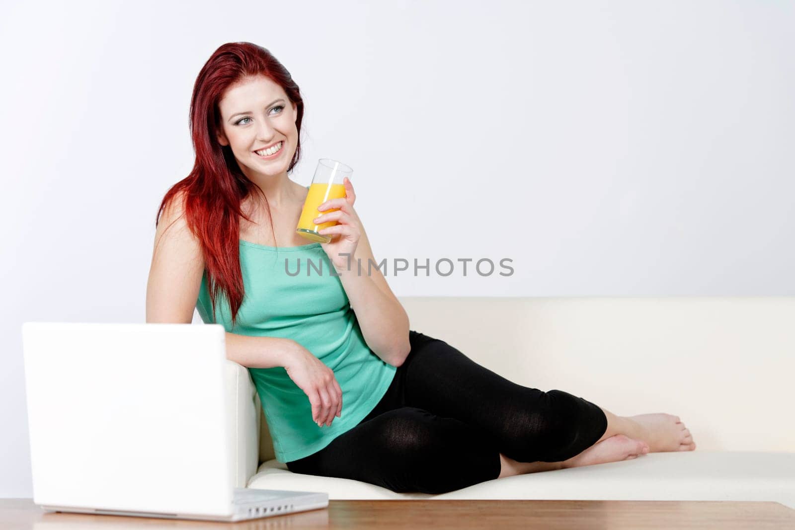 Happy young woman lying on a white sofa at home drinking Orange juice and using a laptop