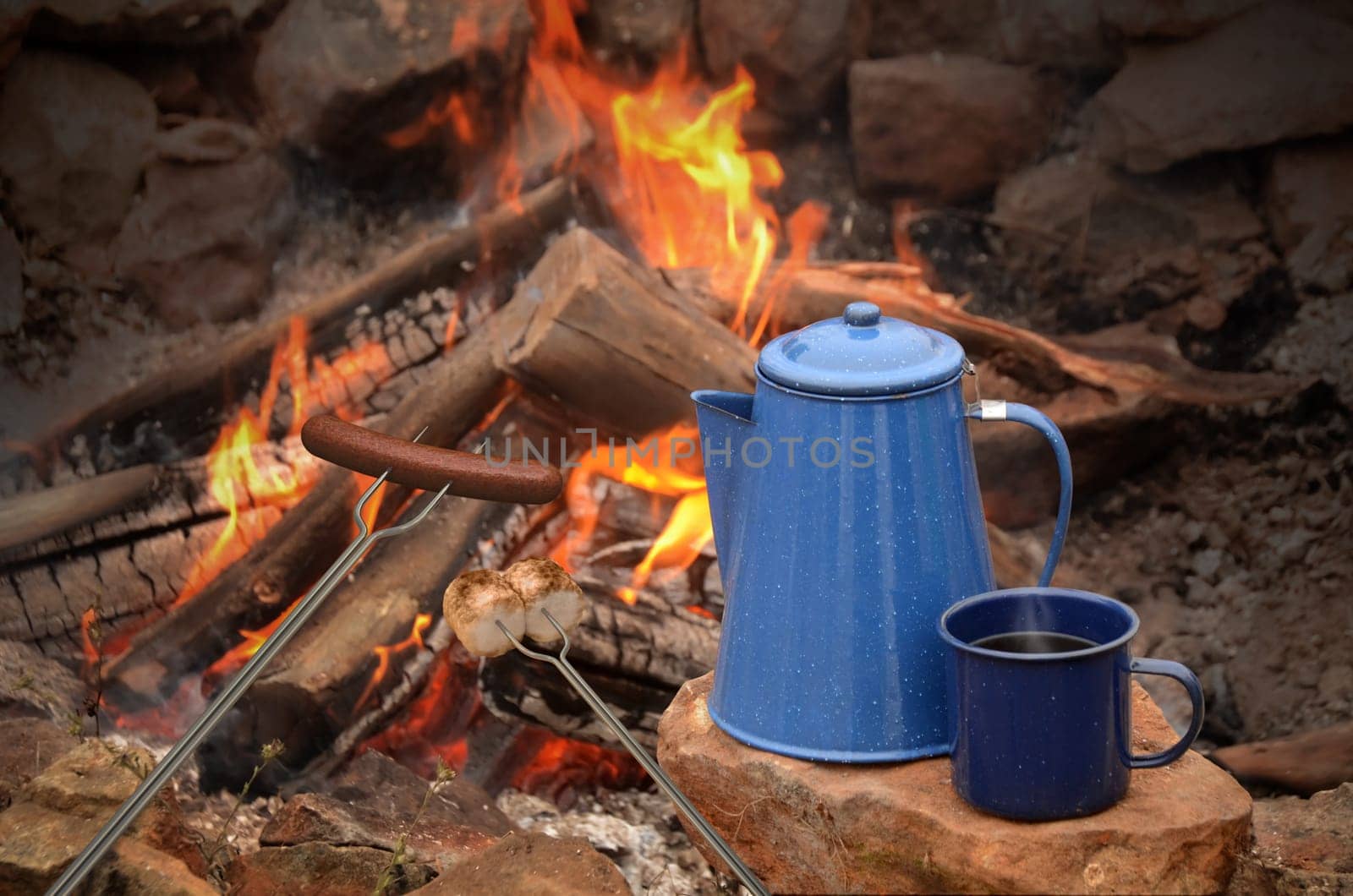 hot dog on a long fork over a fire next to an enamel coffee percolator and mug full of coffee