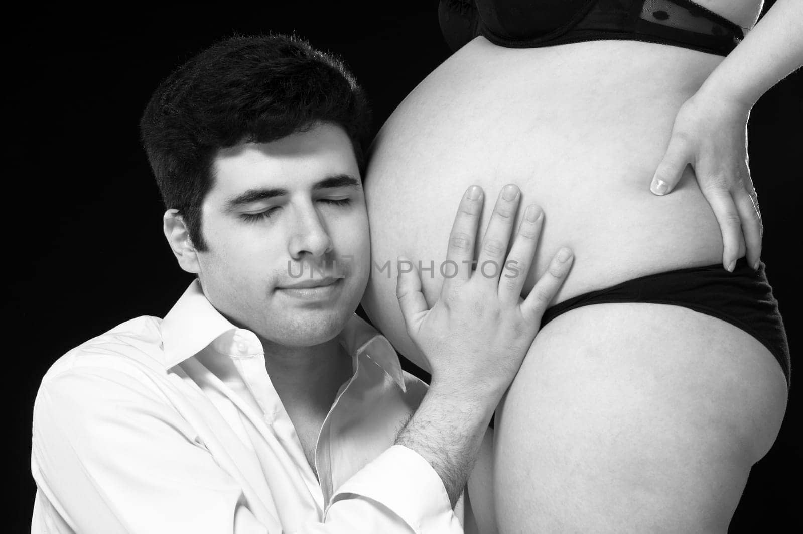 Black and white portrait of young father listening to baby belly with eyes closed.