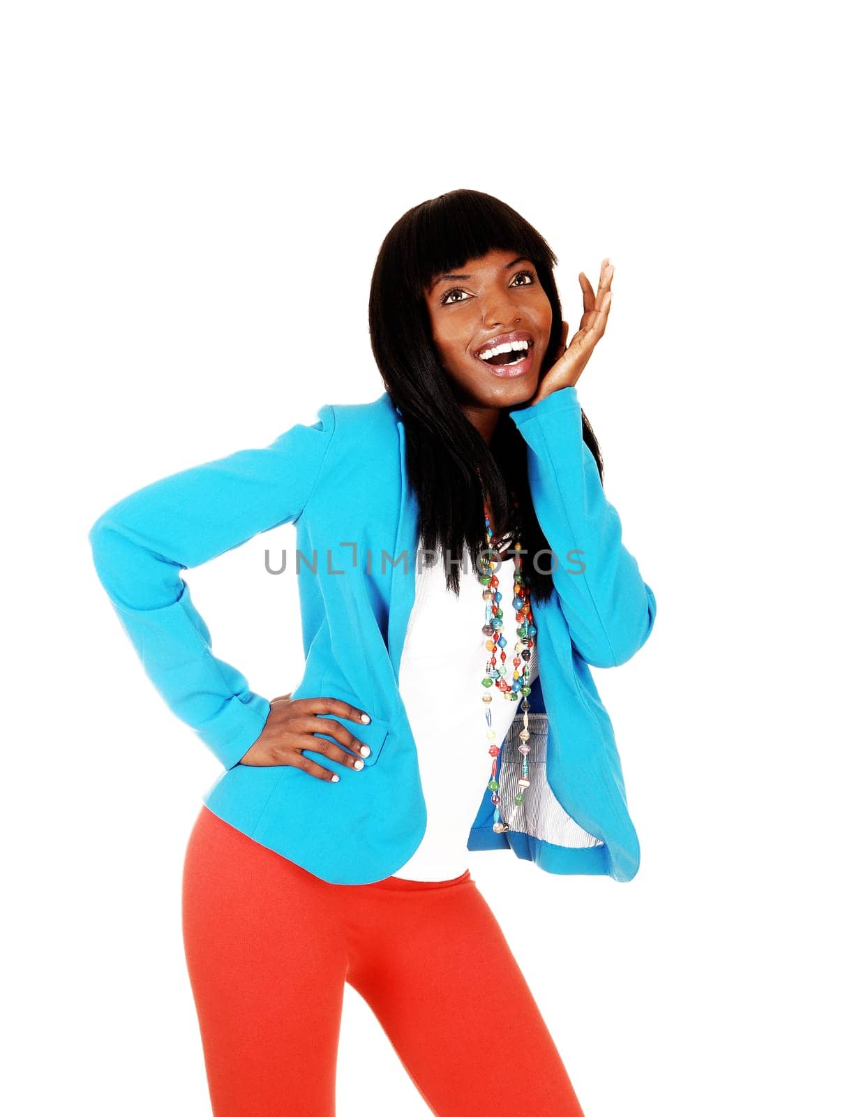 A really happy young afro woman standing for white background in a blue jacket and red tights, holding her hand under her chin.
