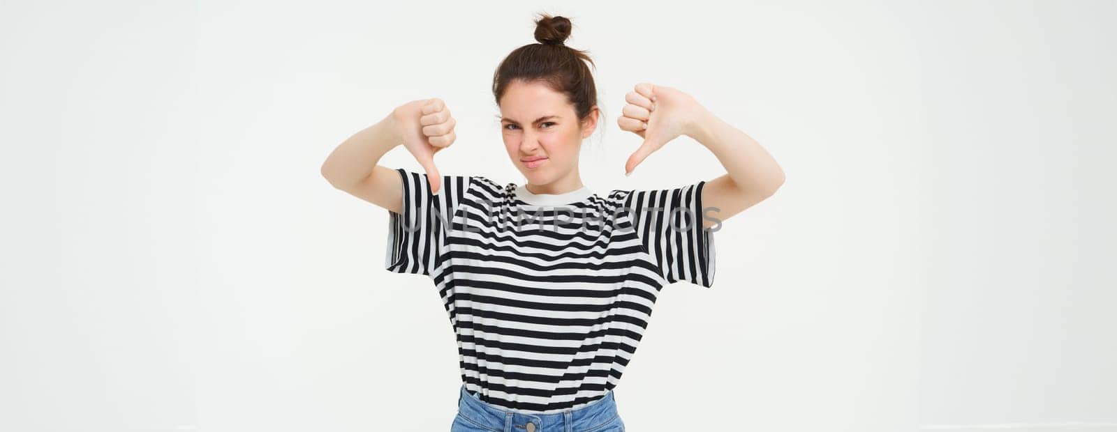 Portrait of disappointed, young modern woman, student shows thumbs down, dislikes smth, disapproves, stands over white background by Benzoix