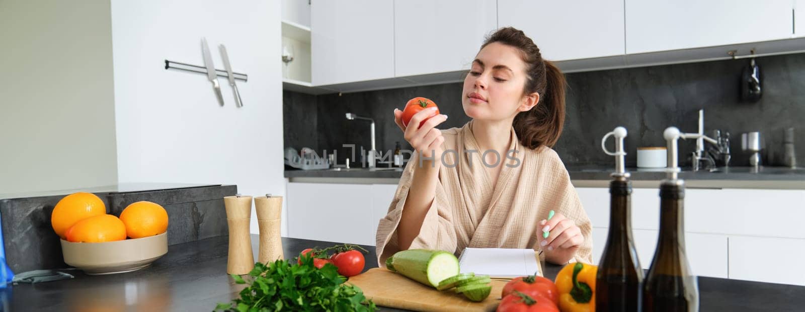 Portrait of beautiful smiling woman, writing her healthy menu, eating tomato while cooking, making grocery list, sitting in the kitchen by Benzoix