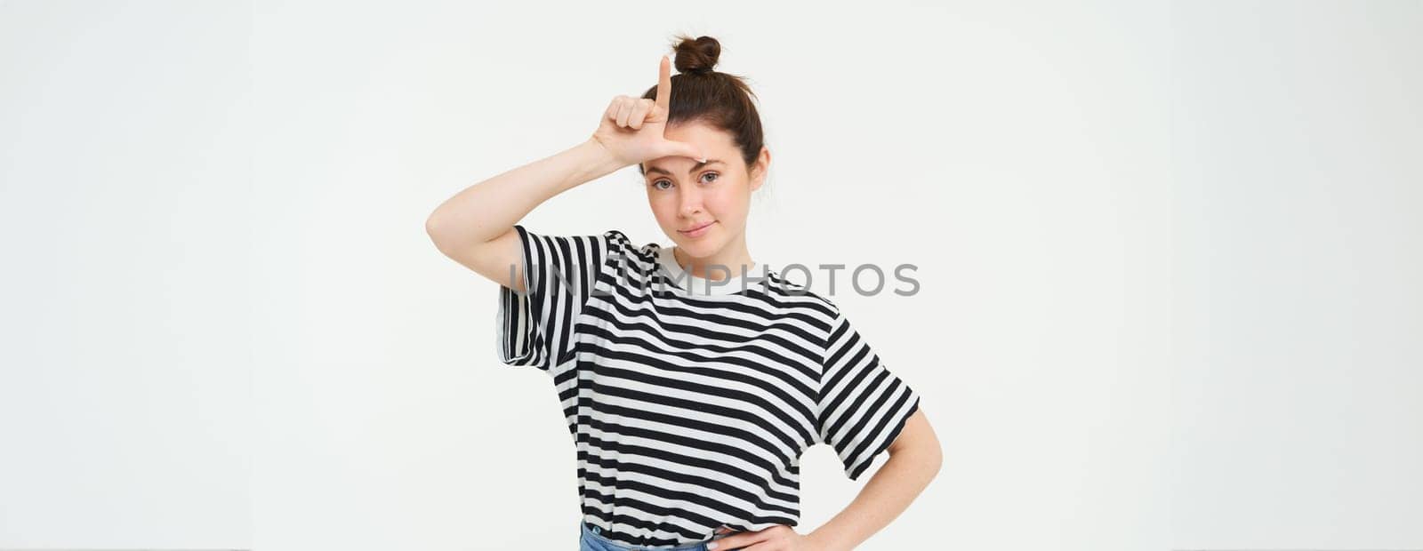Portrait of brunette woman shows l letter on forehead, mocking someone, calling person a loser, standing over white background.