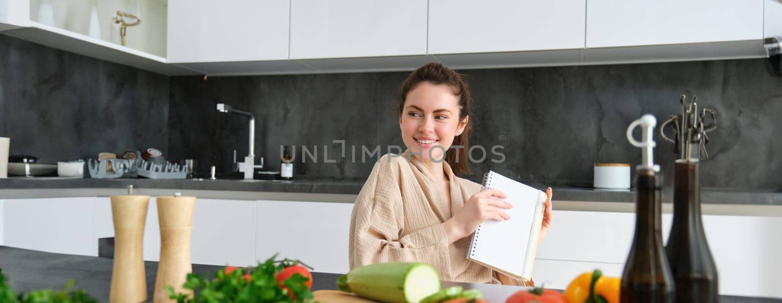 Portrait of woman writing down list of groceries, making notes in recipe, sitting in kitchen near vegetables, preparing dinner menu.