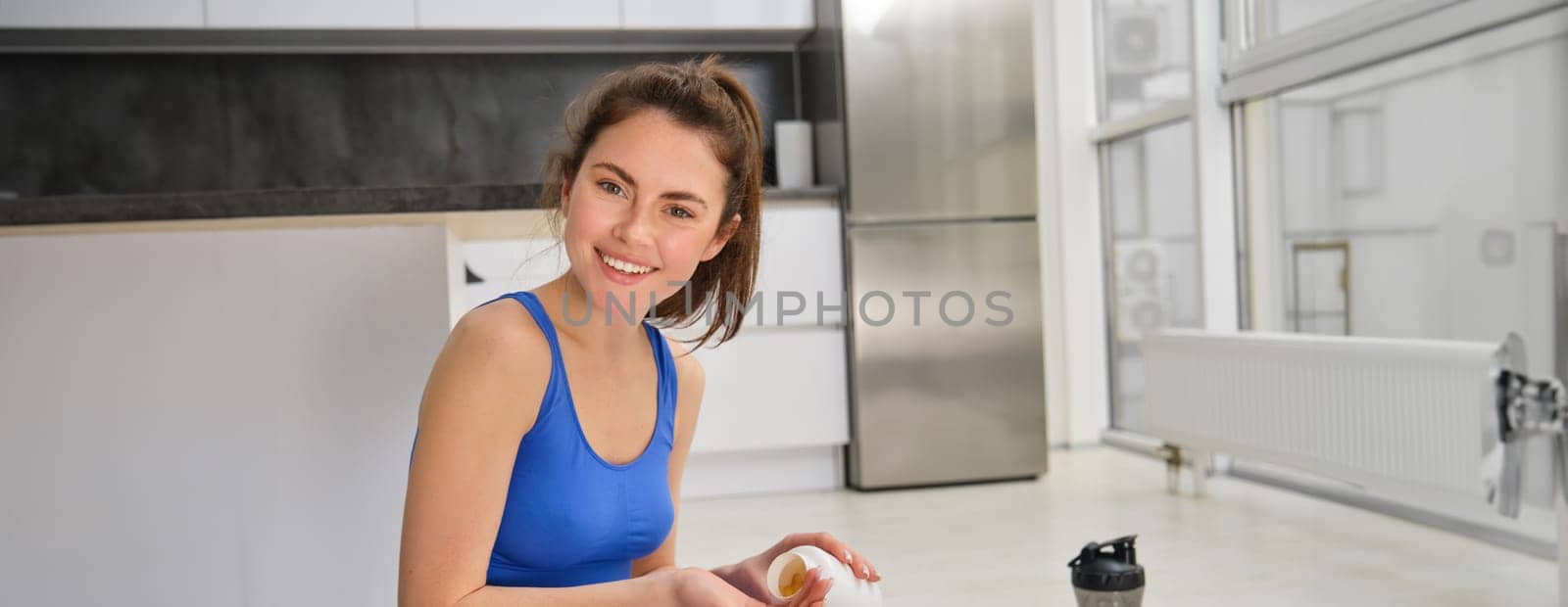 Close up portrait of smiling, healthy young woman, holding tablets, taking vitamin dietary supplements after home fitness workout.