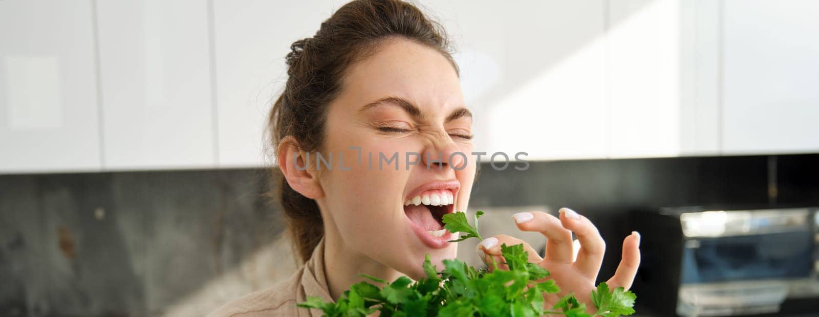 Portrait of attractive woman biting parsley, eating fresh vegetables and herbs in the kitchen, enjoying healthy lifestyle and food.