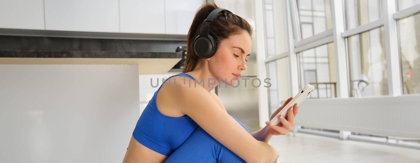 Fitness and wellbeing. Portrait of young woman doing fitness exercises, workout from home, wearing headphones and using smartphone, exercising indoors.
