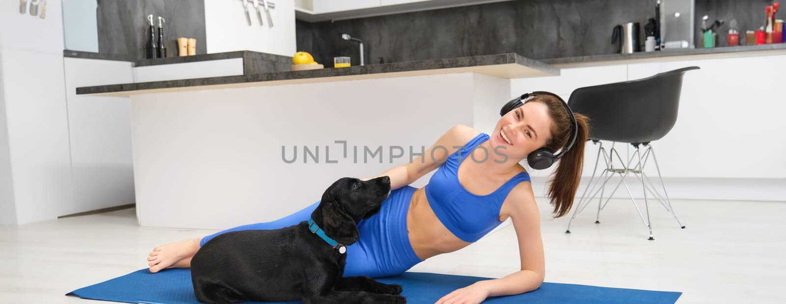 Lifestyle and fitness concept. Happy young woman doing aerobics while her dog wants to play, laughing and smiling, laying on yoga mat by Benzoix