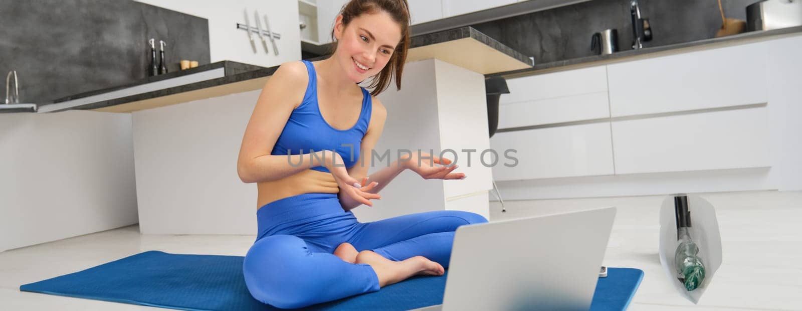 Portrait of fit and healthy, female fitness instructor sitting on floor at home, talking to laptop, teaching online class yoga or workout training by Benzoix