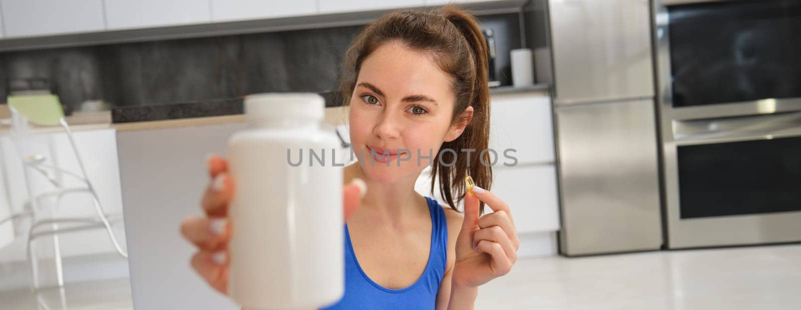 Close up portrait of fitness woman, girl workout from home, showing bottle of vitamins, dietary supplement pills, recommending buds.