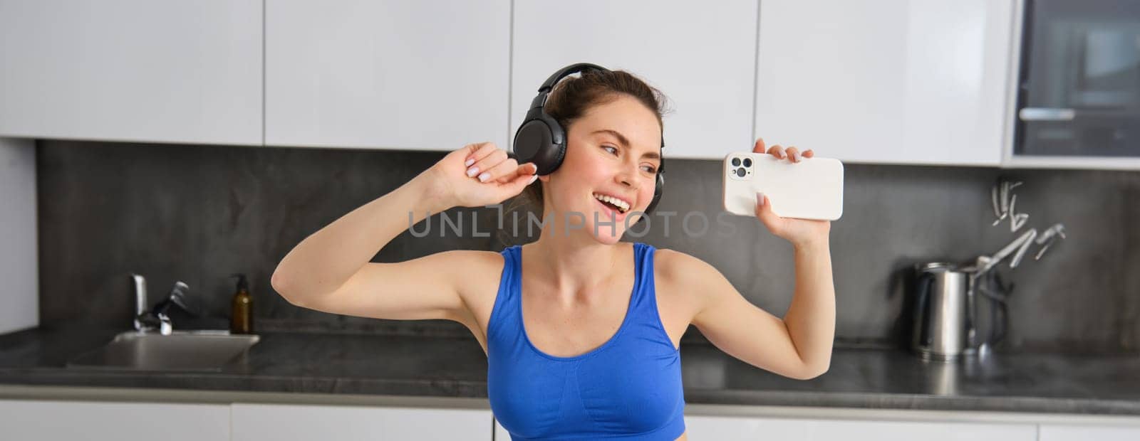 Portrait of beautiful fitness blogger, woman in headphones, listening music and dancing in kitchen, wearing blue leggings and sportsbra.