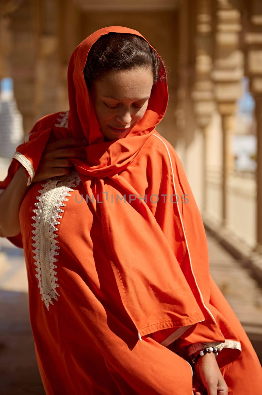 Authentic portrait of Middle Eastern pretty woman with head covered in hijab, dressed in traditional Moroccan Handora dress, posing against beige the mosque marble columns background. People. Religion