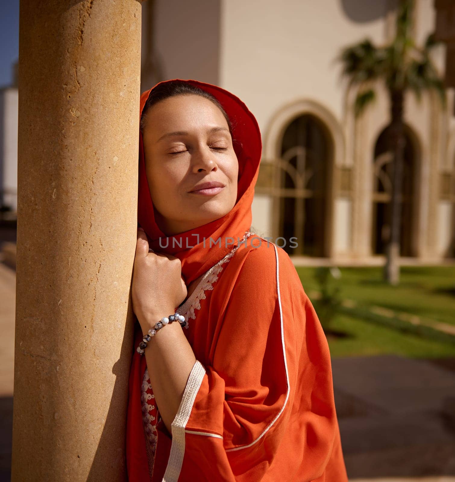 Close-up beauty portrait of gorgeous Arab Muslim woman with her eyes closed, wearing bright orange headscarf and traditional clothes, enjoying beautiful sunrays, leaning on marble column of the mosque