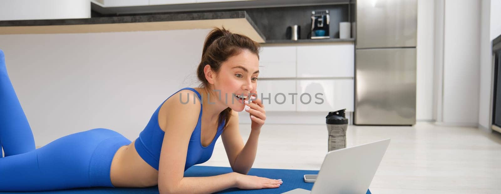 Portrait of smiling, beautiful woman sitting with laptop and watching something, doing fitness exericses from home, workout training in living room.