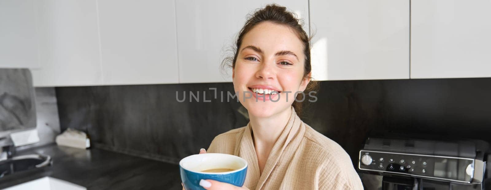 Lifestyle concept. Portrait of happy brunette woman in bathrobe, drinking coffee in the kitchen, having morning cuppa and smiling.