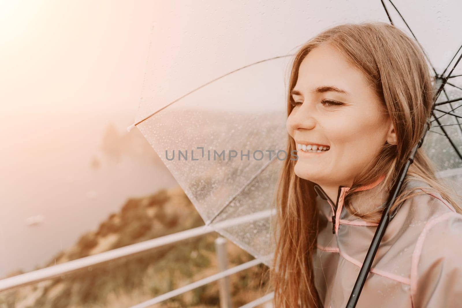 Woman rain umbrella. Happy woman portrait wearing a raincoat with transparent umbrella outdoors on rainy day in park near sea. Girl on the nature on rainy overcast day. by panophotograph