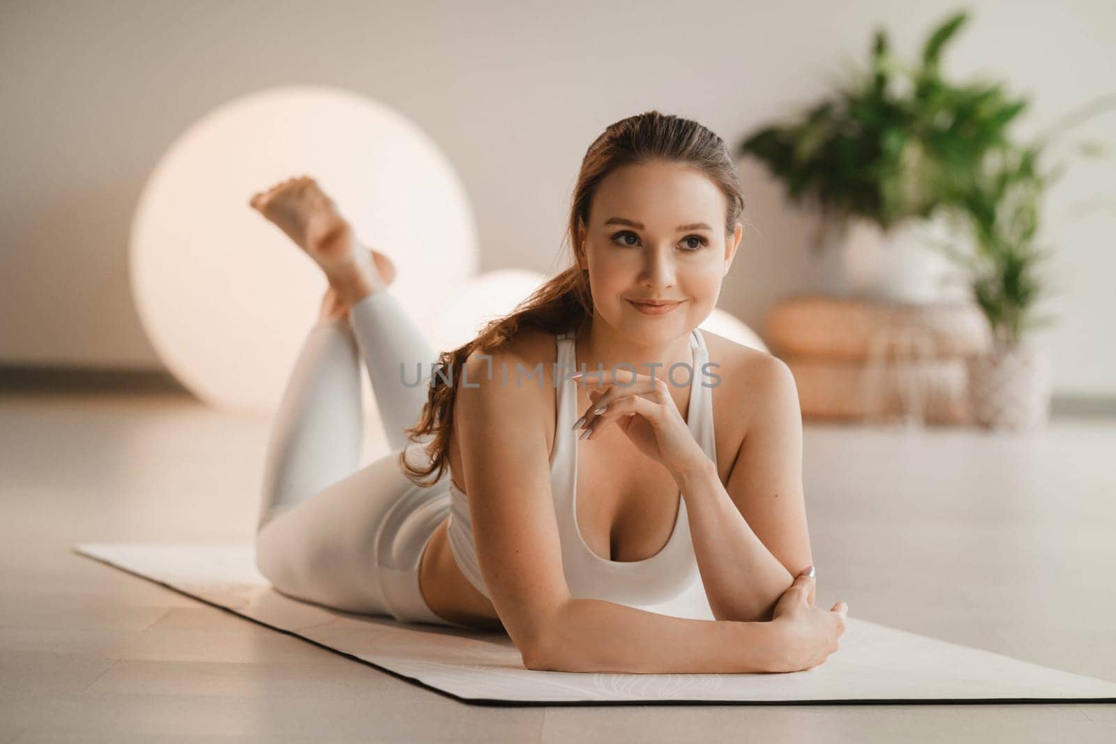 Portrait of a girl in white clothes lying on a mat before doing yoga indoors by Lobachad