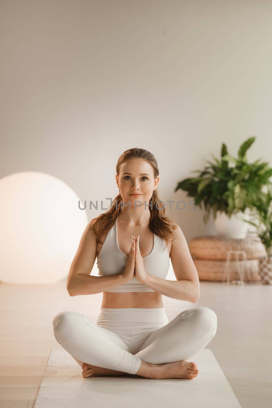 Portrait of a girl in white clothes sitting in a lotus position on a mat at an indoor yoga class.