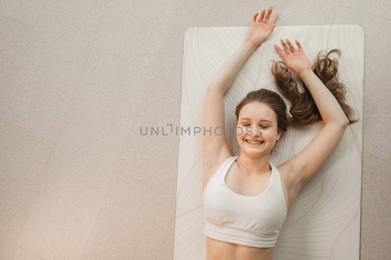 A girl in white clothes lies on a rug and laughs before yoga classes indoors by Lobachad