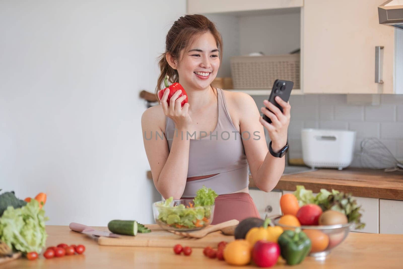 Beautiful young woman in exercise clothes having fun in a cute kitchen at home. Using the phone to study information And prepare vegan fruit salad dressings, fruit shakes, or healthy smoothies..