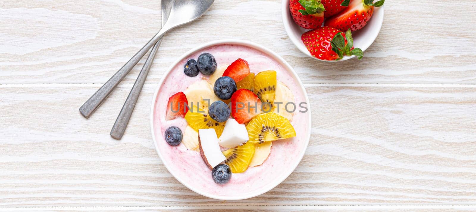 Healthy breakfast or dessert yogurt bowl with fresh banana, strawberry, blueberry, cocos, kiwi top view, rustic wooden white background with spoon by its_al_dente