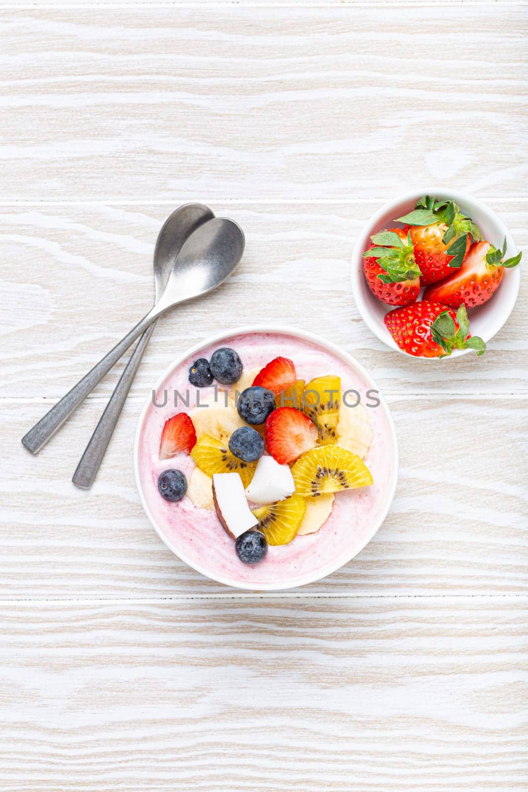 Healthy breakfast or dessert yogurt bowl with fresh banana, strawberry, blueberry, cocos, kiwi top view on rustic wooden white background with spoon