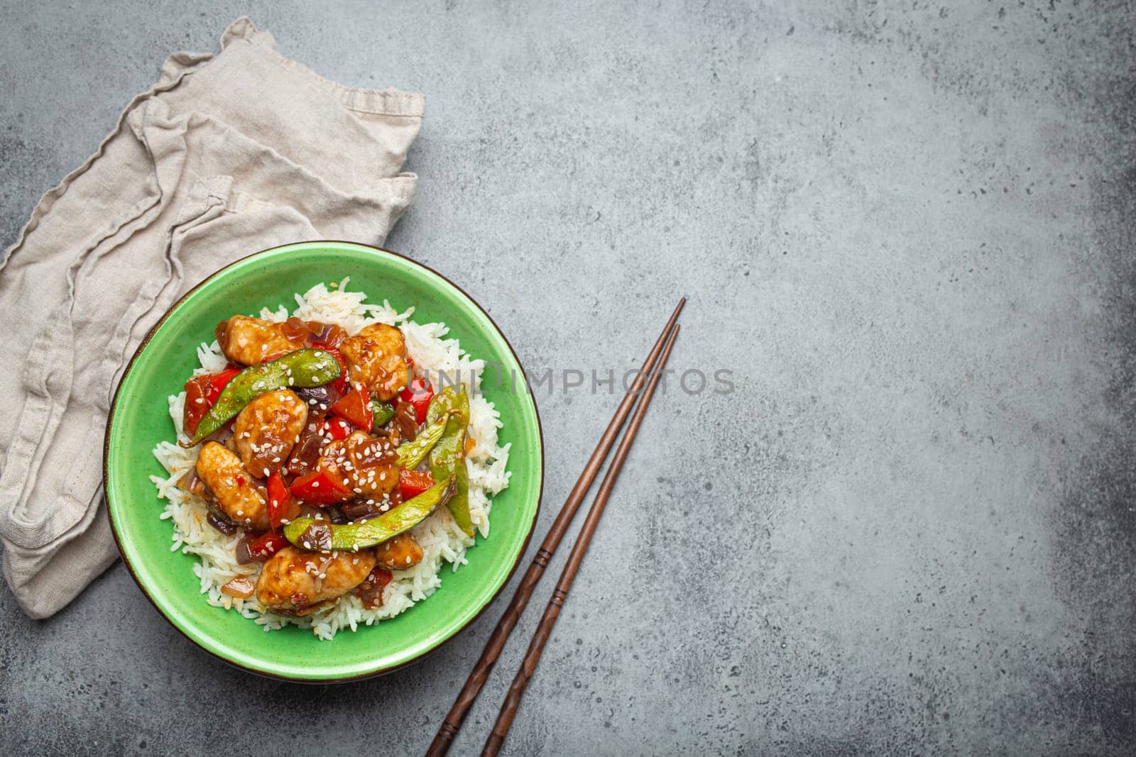 Asian sweet and sour sticky chicken with vegetables stir-fry and rice in ceramic bowl with chopsticks top view on gray rustic stone background, traditional Asian dish. Copy space by its_al_dente