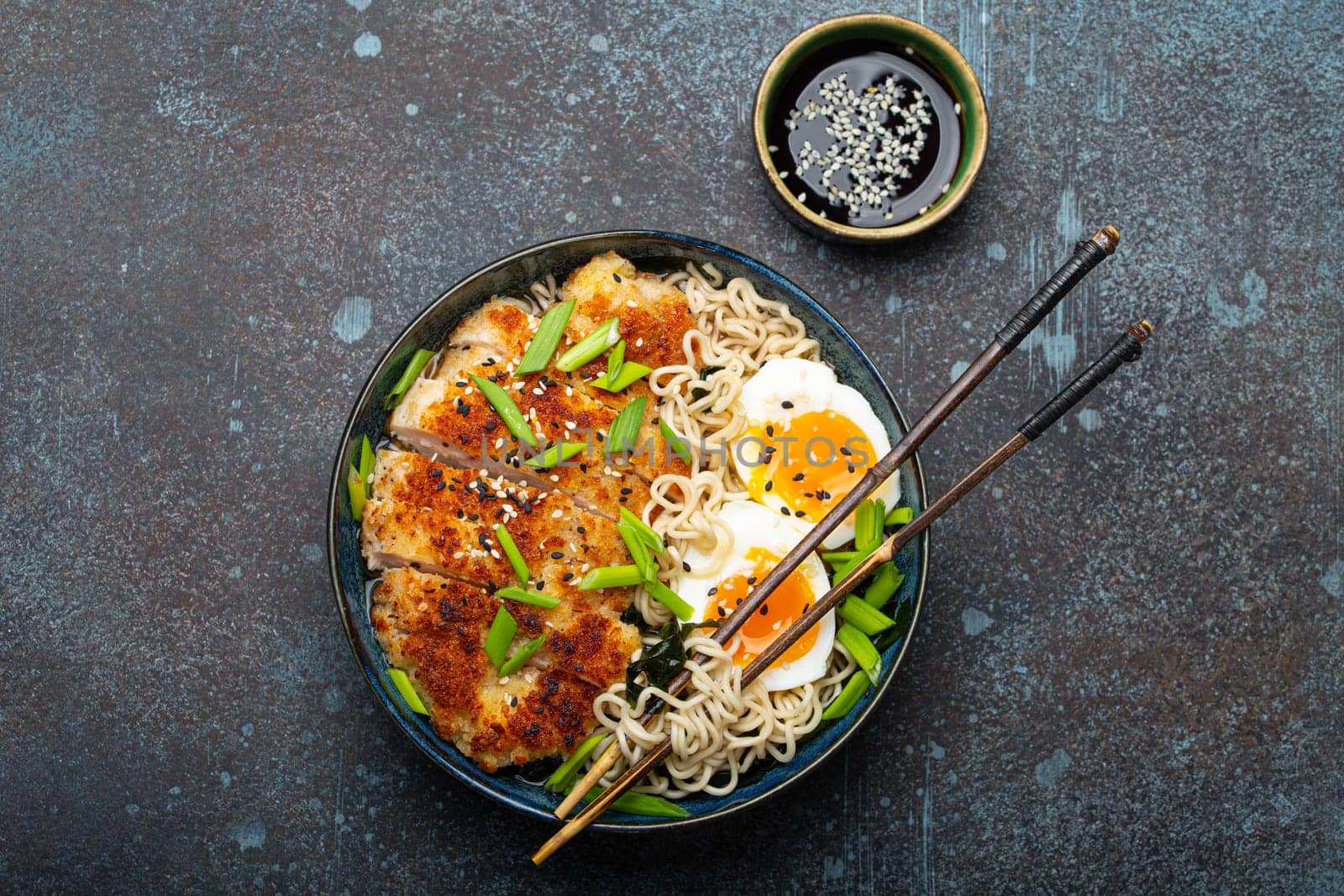 Asian noodles ramen soup with deep fried panko chicken fillet and boiled eggs in ceramic bowl with chop sticks, soy sauce on stone rustic background top view by its_al_dente