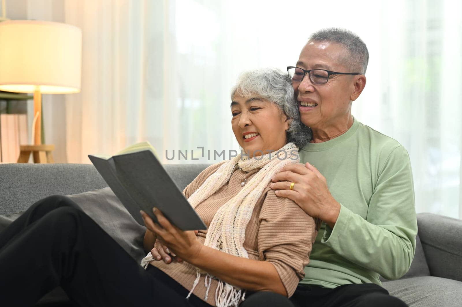 Loving senior couple embracing and reading book on sofa while spending time together at home.