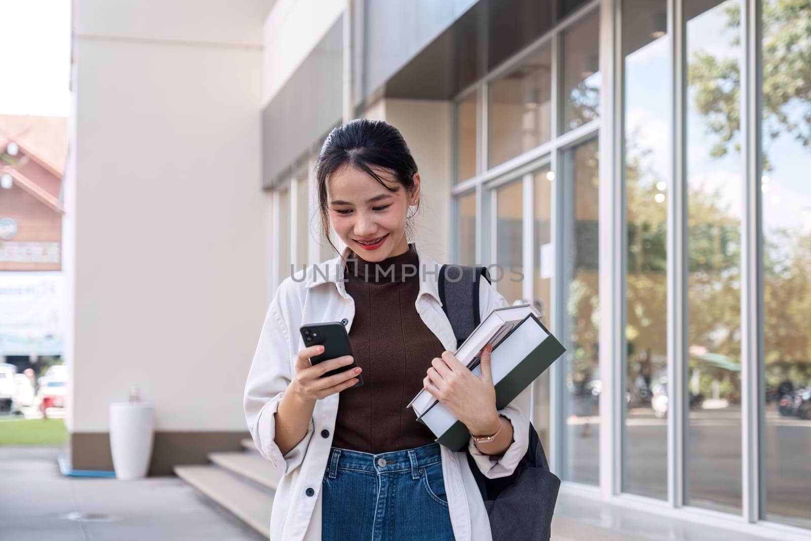 Attractive young Asian female student stands holding a book and using her phone to make an appointment with a college friend..