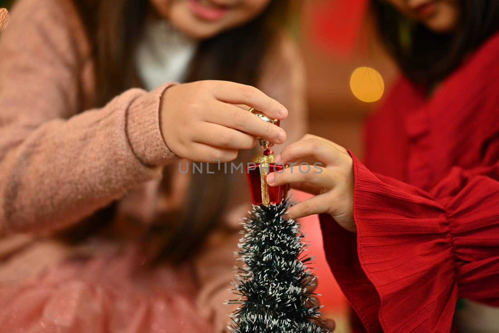 Hands of little children wearing warm winter clothes decorating small Christmas tree in living room.