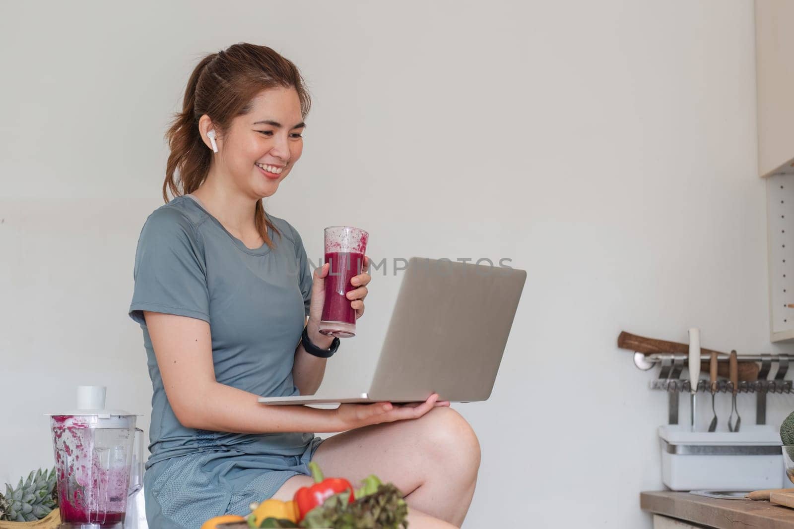 Beautiful young woman in exercise clothes having fun in a cute kitchen at home. Using your laptop to prepare vegan fruit salad dressing, fruit shakes, or healthy smoothies..