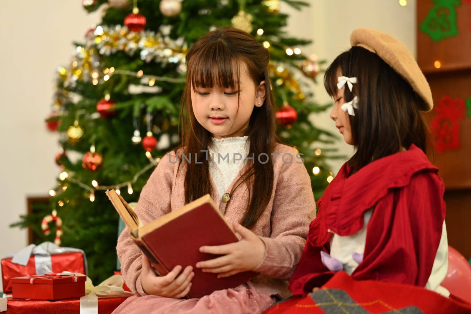 Happy little siblings reading a storybook together in cozy living room decorated for Christmas.